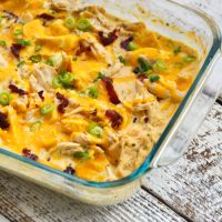 fully baked chicken topped with shredded cheddar cheese, bacon, and scallions in a glass baking dish