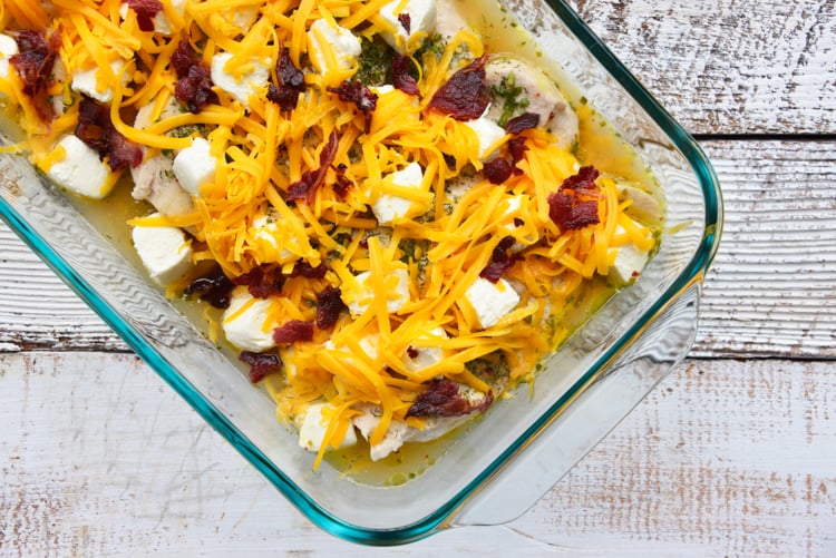 half baked chicken topped with cream cheese cubes and shredded cheese in a glass baking dish