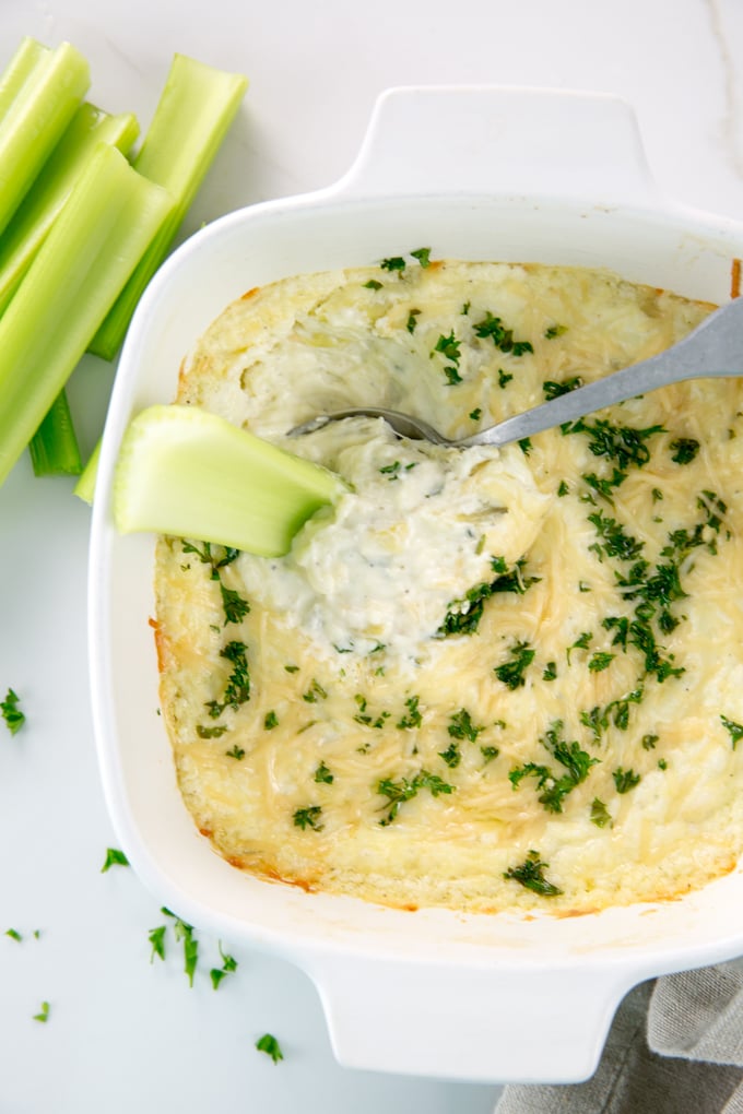 Vertical image of Baked Creamy garlic parmesan hot artichoke dip in a white square casserole dish with celery on the side and in the bowl
