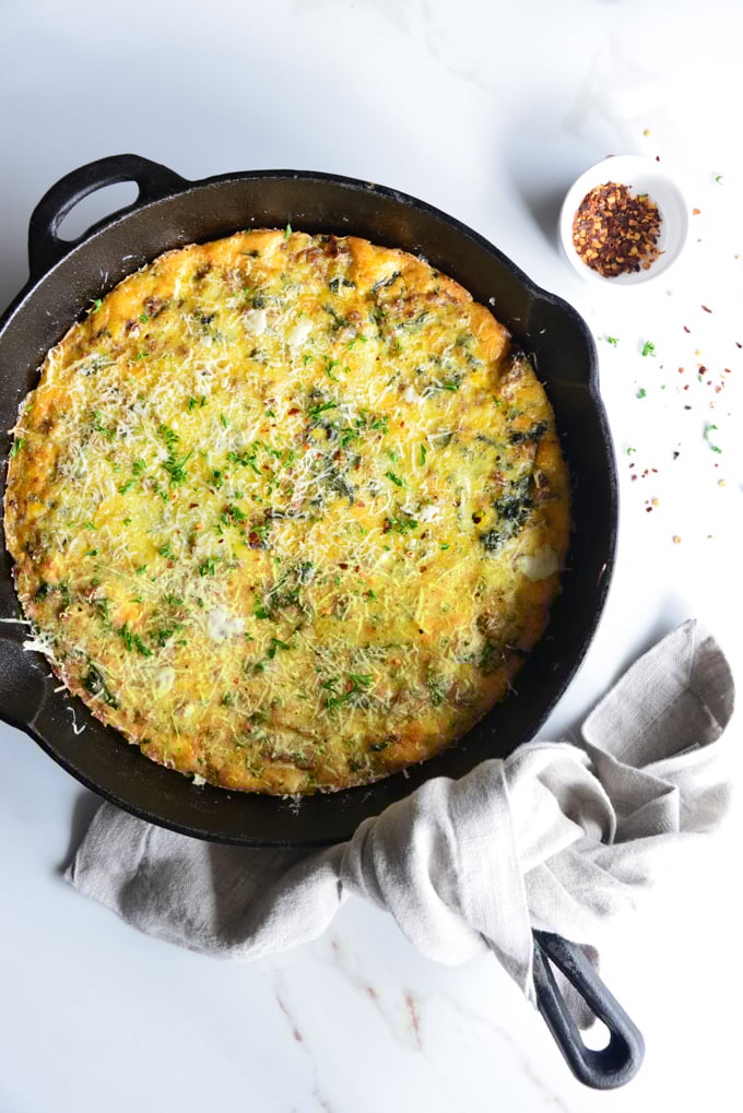 vertical image of a baked frittata in a cast iron skillet with a cloth napkin and red pepper flakes in a small dish