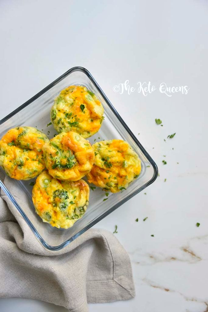 Overhead close up vertical image of broccoli and cheddar egg muffins in a glass storage container on a white background