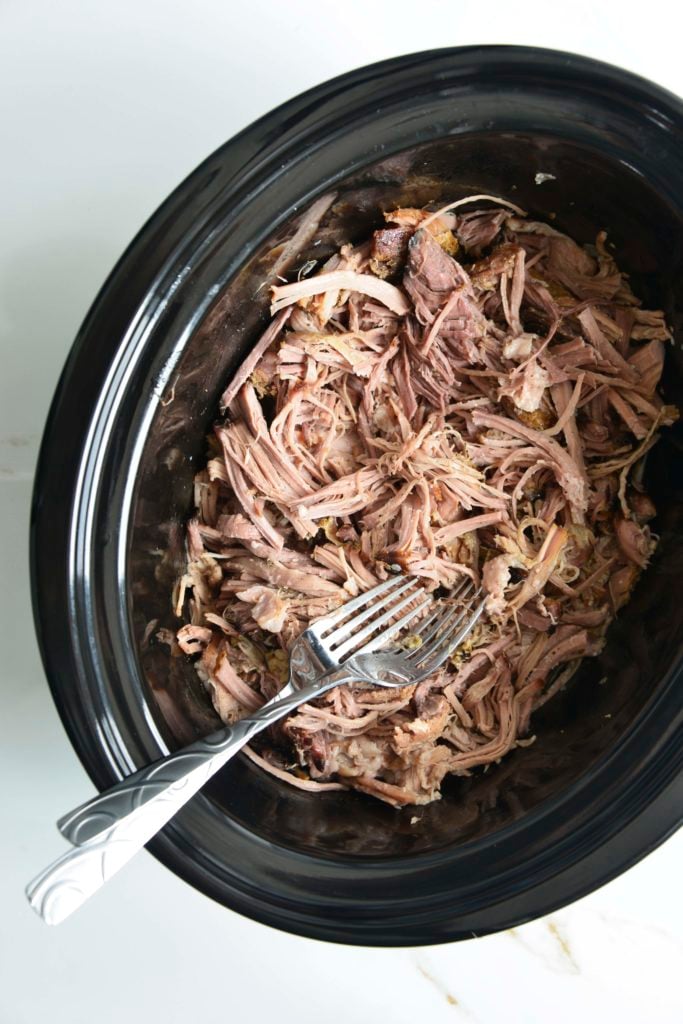 shredded pork in a black slow cooker with 2 forks