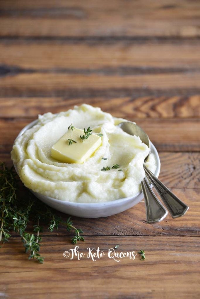 Pinterst image for low carb butter garlic mashed cauliflower Vertical image of 3/4 shot of mashed cauliflower in a bowl with butter and fresh thyme on top with 2 spoons