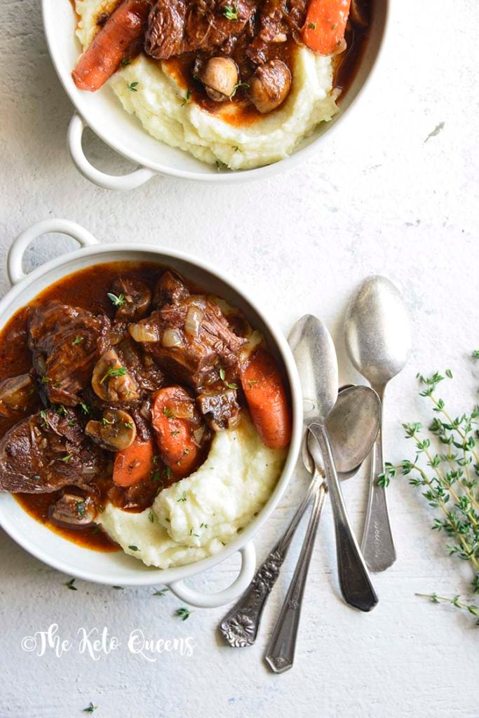 vertical overhead image of low carb instant pot beef stew in a white bowl with 3 spoons and fresh herbs on a white background