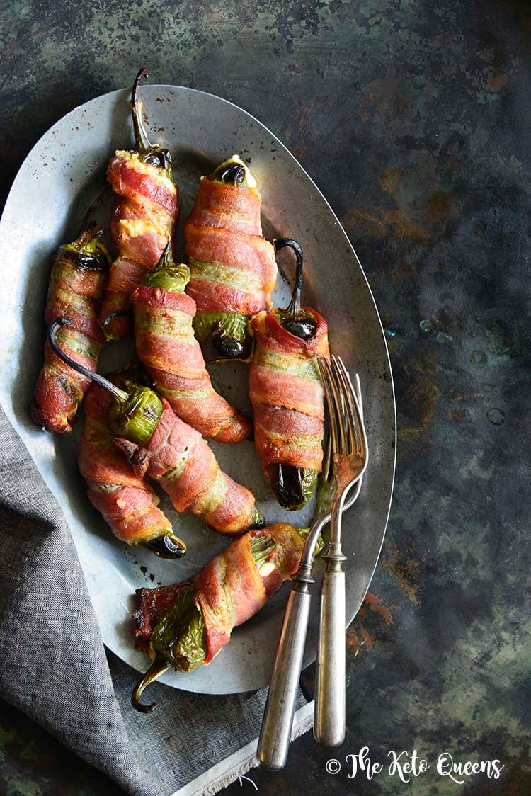 Our Bacon Wrapped Jalapeno Poppers are a keto lovers dream! They're smokey, stuffed with 2 kinds of cheese and wrapped in delicious bacon! Need we say more? Keto Bacon Wrapped Jalapeno Poppers on a silver tray with 2 forks on a dark background