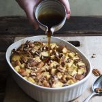 vertical image of maple pecan french toast casserole in a white casserole dish, with a spoon on a wood background with low carb syrup being poured onto it
