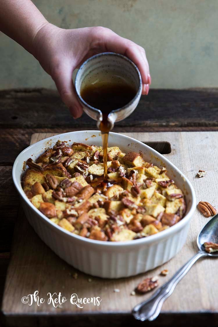vertical image of maple pecan french toast casserole in a white casserole dish, with a spoon on a wood background with low carb syrup being poured onto it